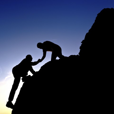 A Silhouette of One Person Helping Another Climb Up a Mountain