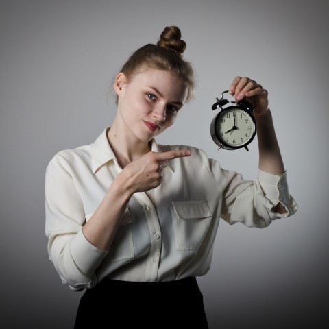 A Woman in Front of a Gray Wall Holding Up an Alarm Clock and Pointing at It