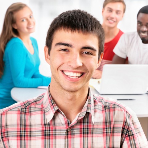 A Guy in a Checkered-shirt Smiling into the Camera with Peers behind Him also Smiling