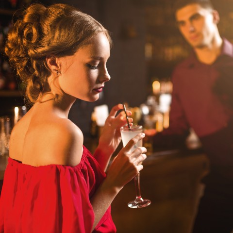 Young Woman in a Red Dress Holding a Drink with a Young Man in the Background Looking at Her